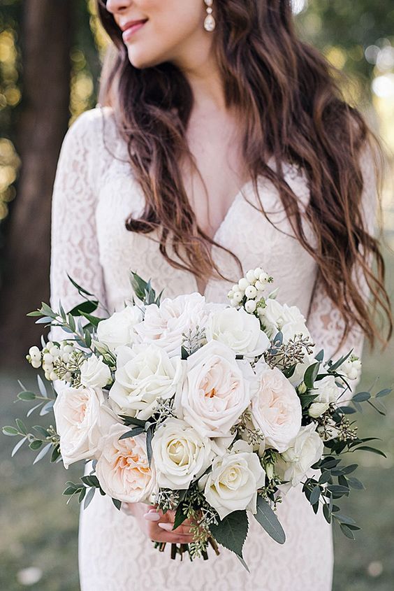 Mariée dans une robe blanche avec des longues manches et un décolleté en V avec un bouquet de roses banches et des feuillages 